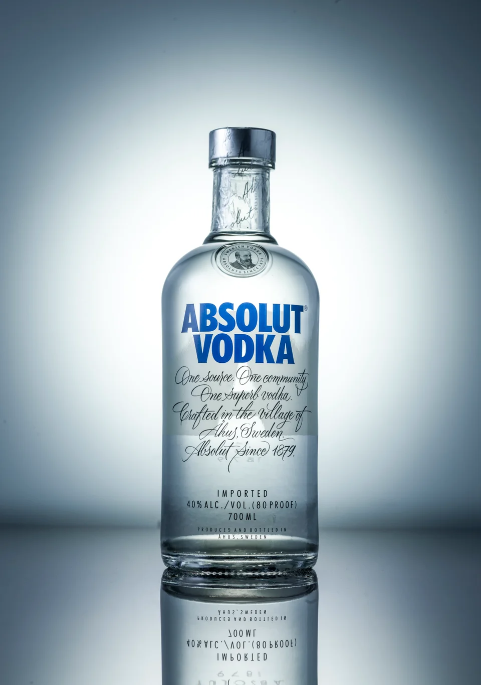 A glass Absolut bottle lit against a cool gradient background, reflected on the surface below.