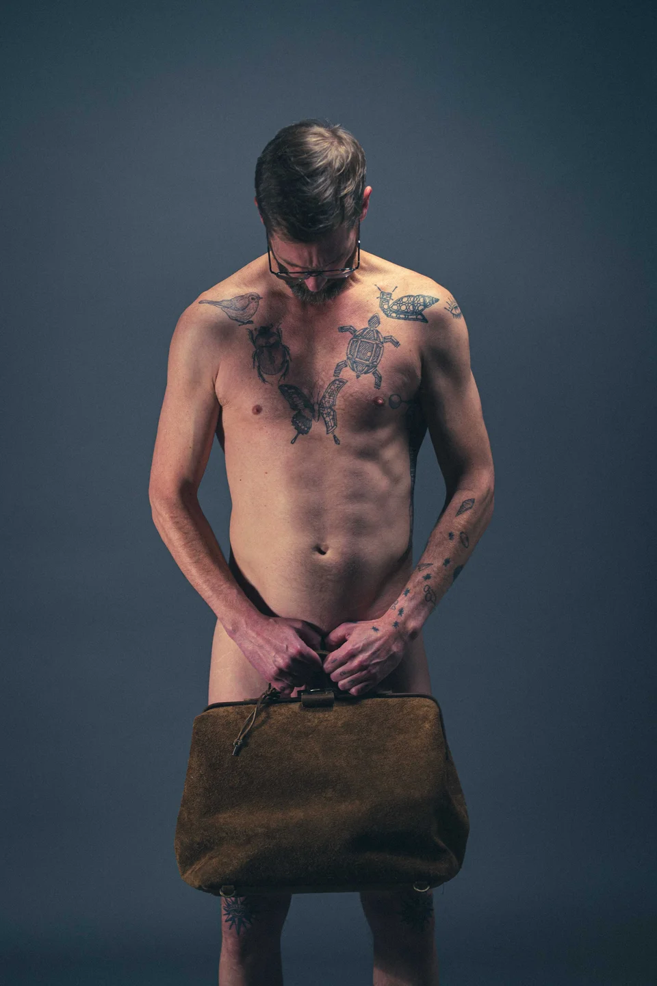 A shirtless person looking down while holding a brown bag in a dark studio.